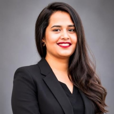Professional headshot of a woman with long dark hair wearing a black blazer and red lipstick, smiling at camera against gray background