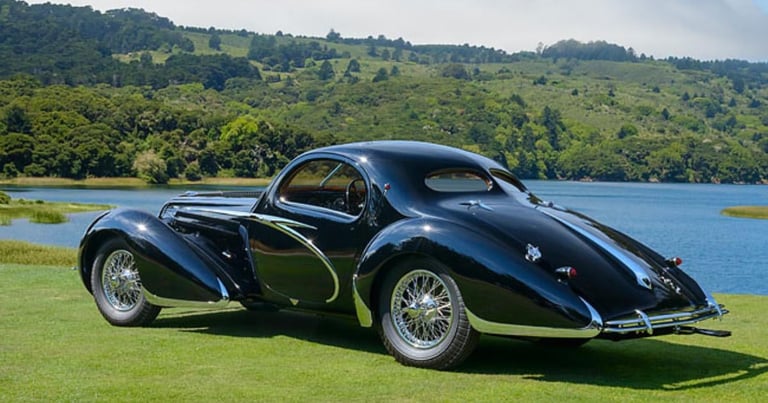 Black vintage sports car with open door parked on grass near blue water and forested hills