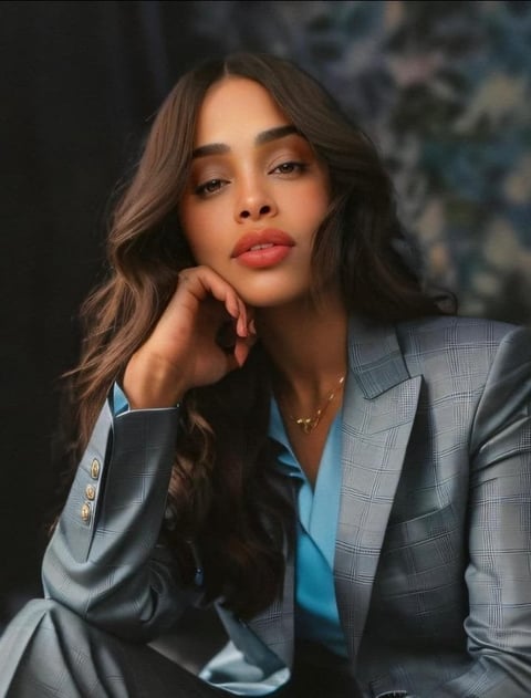Woman in gray plaid blazer and blue shirt resting hand on face, looking at camera against blurred background