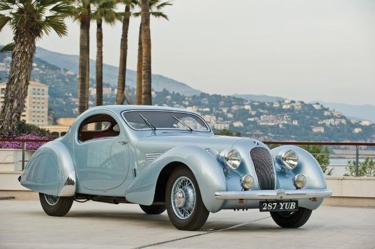 Light blue vintage coupe car parked by palm trees with hillside houses in background