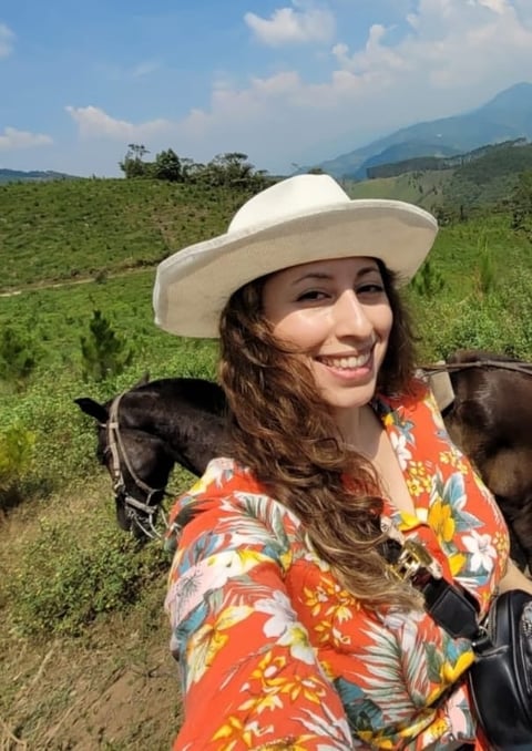 Woman in white cowboy hat and colorful orange floral shirt smiling with horses on rural mountain farm landscape
