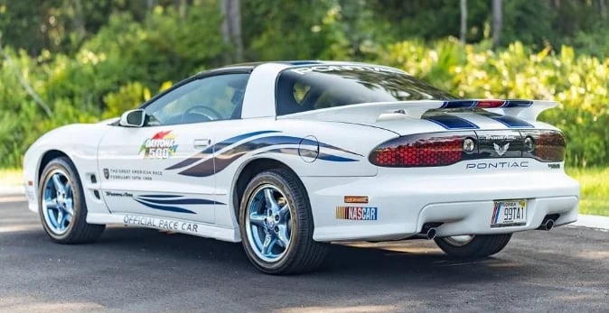 White Pontiac Grand Am with blue racing stripes parked on a road with green foliage in background