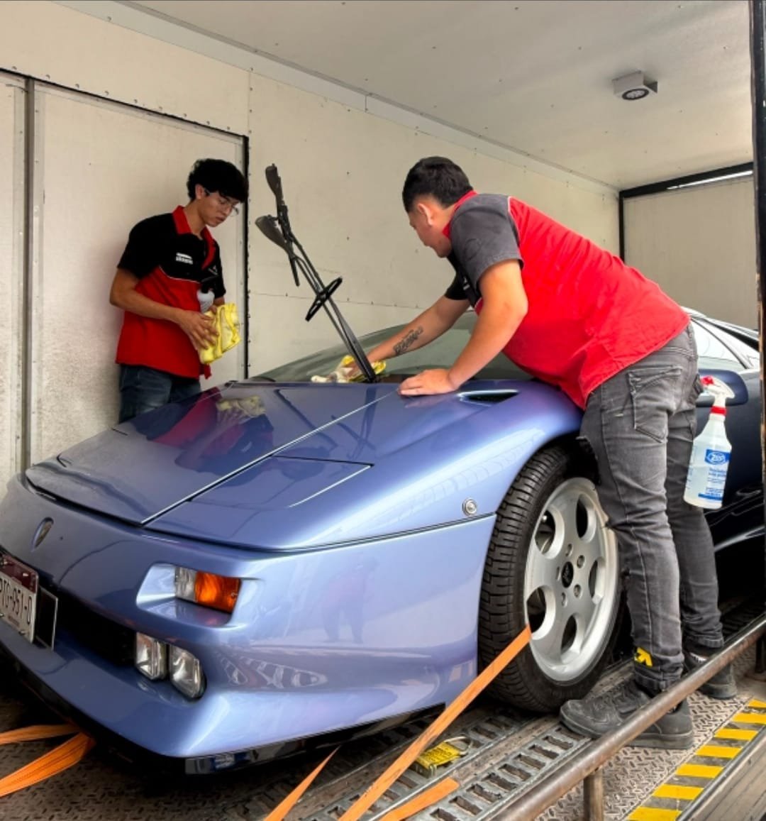 Two technicians in red shirts washing a blue sports car in an indoor garage bay with equipment